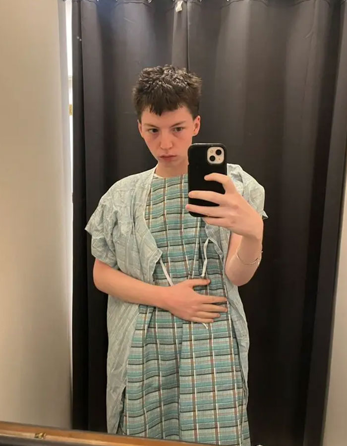 Young person in hospital gown taking a mirror selfie, representing model pushed to transition at 15 now detransitioning. Young person in hospital gown taking a mirror selfie, representing model pushed to transition at 15 now detransitioning.