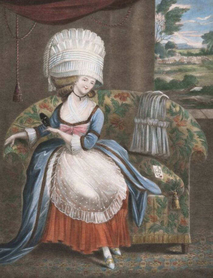 18th-century courtesan dressed in elaborate gown and headdress, seated on a floral-patterned sofa indoors.