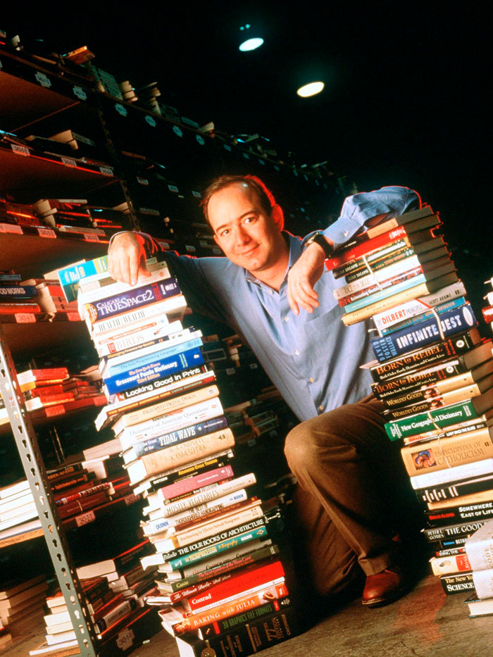 Man resembling Jeff Bezos with hair, leaning against stacks of books in a dimly lit room, ditching his signature bald look. Man resembling Jeff Bezos with hair, leaning against stacks of books in a dimly lit room, ditching his signature bald look.
