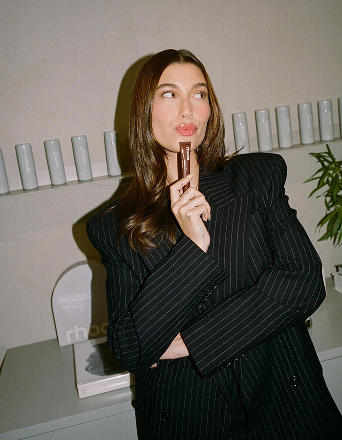 Hailey Bieber posing with Rhode product, wearing a black pinstripe blazer in a minimalist modern setting. Hailey Bieber posing with Rhode product, wearing a black pinstripe blazer in a minimalist modern setting.