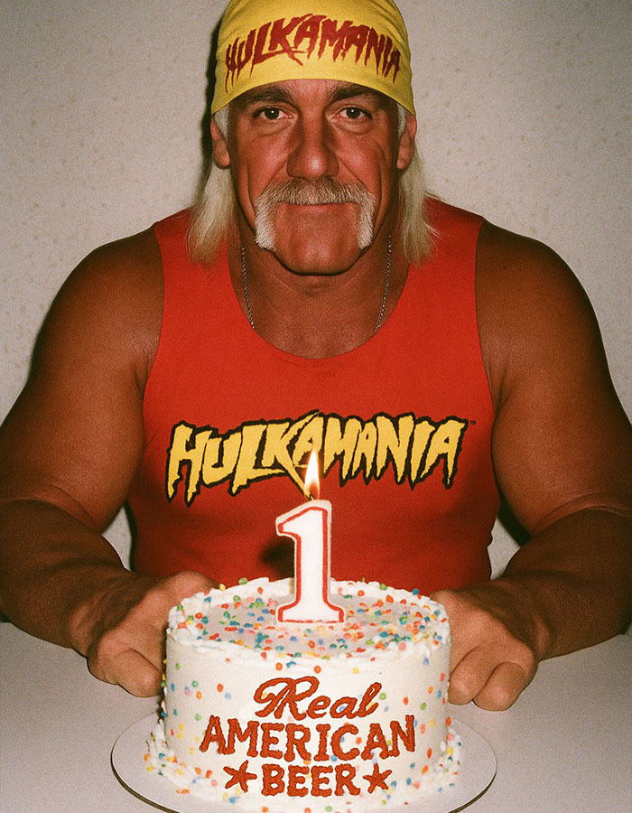Hulk Hogan wearing red Hulkamania shirt and yellow bandana, holding a cake with a lit number one candle. Hulk Hogan wearing red Hulkamania shirt and yellow bandana, holding a cake with a lit number one candle.
