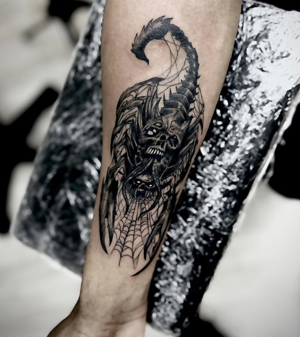 Black and gray tattoo of a detailed scorpion with skull faces and spider webs on a personu2019s forearm.