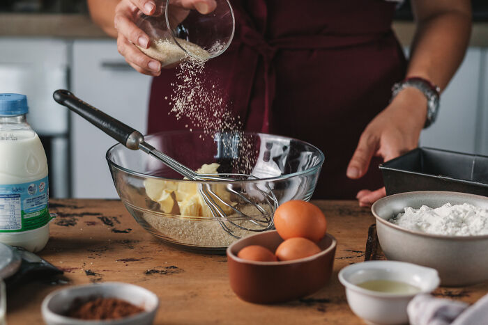 Person pouring sugar into a bowl with butter and whisk, preparing ingredients to bake a childhood cake recipe. Person pouring sugar into a bowl with butter and whisk, preparing ingredients to bake a childhood cake recipe.