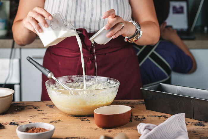 Person wearing apron pouring milk and another liquid into a bowl, baking childhood cake as a family remedy after hospital trip. Person wearing apron pouring milk and another liquid into a bowl, baking childhood cake as a family remedy after hospital trip.