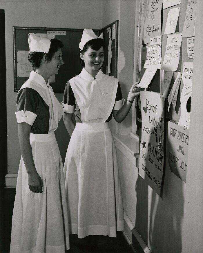 Two vintage nurses in classic uniforms smiling and reading notices on a bulletin board in a hospital hallway.