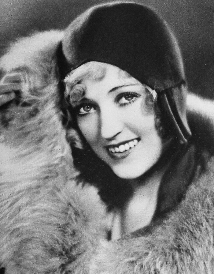 Black and white portrait of a smiling woman wearing a vintage hat and fur coat, showcasing timeless beauty from a century ago.