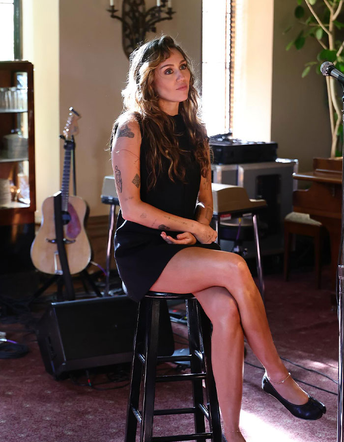 Miley Cyrus sitting on a stool in a black dress, with tattoos visible, during a music studio interview session. Miley Cyrus sitting on a stool in a black dress, with tattoos visible, during a music studio interview session.