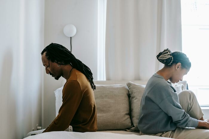 Man and woman sitting back to back on bed, appearing upset and distant in a neutral-toned bedroom setting.
