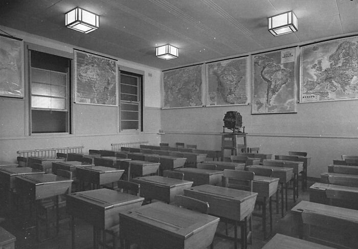 Empty classroom with wooden desks and world maps on walls in a school during World War II time period.