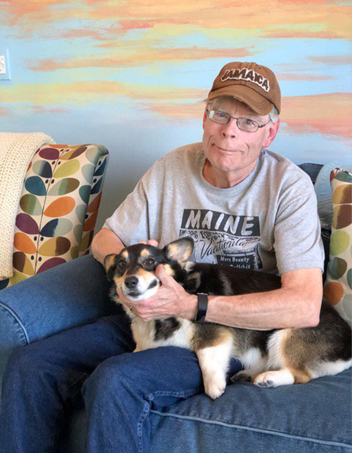 Stephen King sitting on sofa with dog, wearing a Maine t-shirt and Jamaica cap amid Epstein List conspiracy theories. Stephen King sitting on sofa with dog, wearing a Maine t-shirt and Jamaica cap amid Epstein List conspiracy theories.