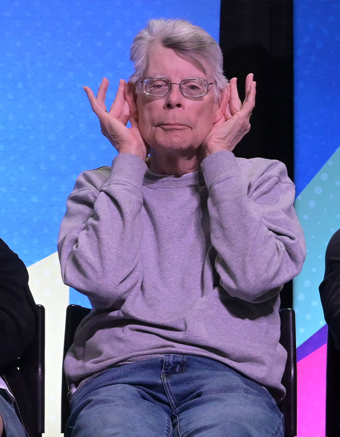 Stephen King wearing glasses and a gray sweatshirt, making a playful gesture with hands near ears on stage. Stephen King wearing glasses and a gray sweatshirt, making a playful gesture with hands near ears on stage.