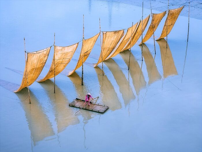 Fisherman navigating a small wooden raft near traditional yellow fishing nets reflecting on calm Asian waters.