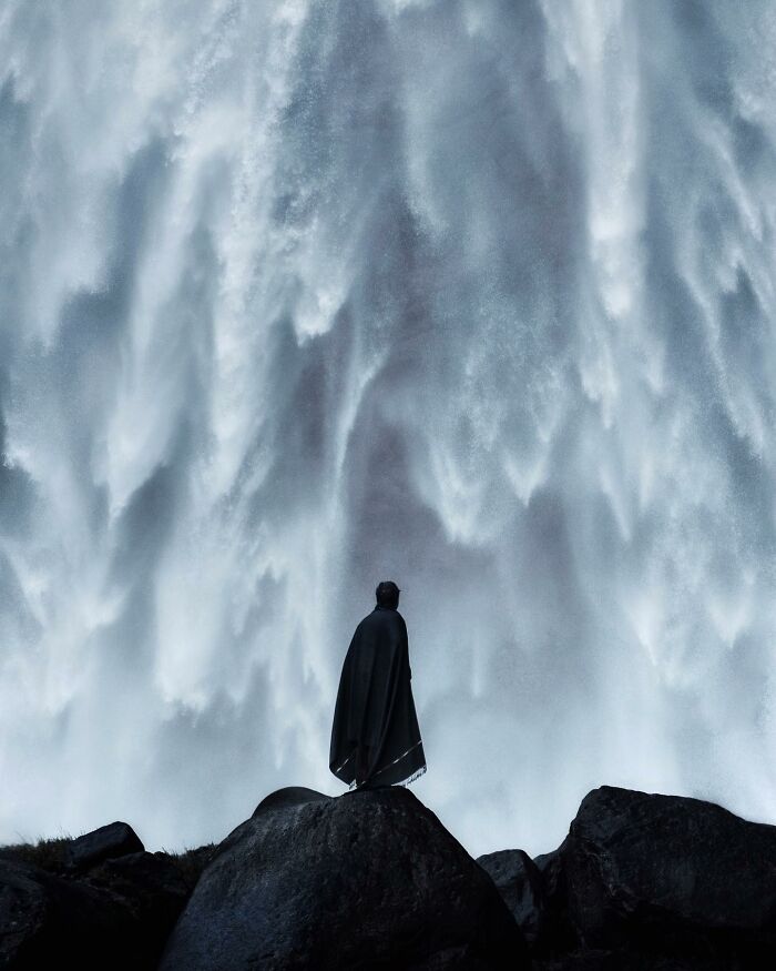 Person standing on rock in front of a massive cascading waterfall in a breathtaking landscape shot resembling a fantasy scene.