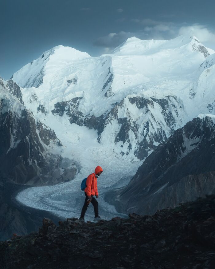 Hiker in orange jacket stands on rocky terrain with breathtaking landscape of snow-covered mountains and glacier in the background.