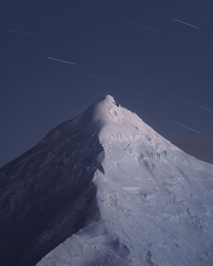 Snow-covered mountain peak at night under star trails, a breathtaking landscape shot resembling a fantasy movie scene.