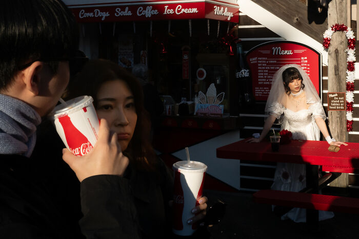 Couple drinking soda near a woman in a wedding dress at a red picnic table, captured in a coincidental public moment.