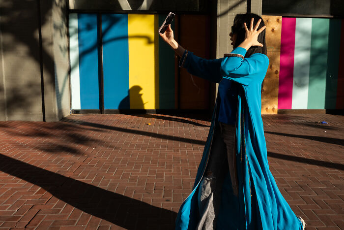 Woman in a blue coat taking a selfie in public with colorful wall panels and shadows, captured in a coincidental moment.