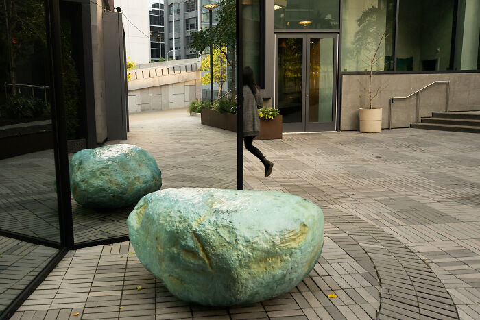 Large blue-green rock near reflective glass wall in a public urban space capturing coincidental moments by photographer.