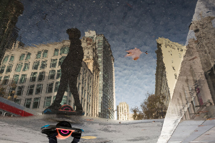 Reflection of a person and buildings in a puddle, capturing a coincidental moment in public by a photographer.
