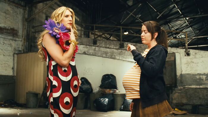 Two women in a gritty warehouse scene from one of the worst movies, one wearing a striped dress and the other with a fake pregnancy belly.