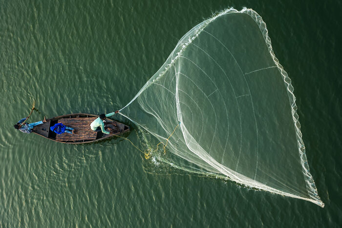 Two fishermen on a boat casting a large net over green water in spectacular mangrove photography awards image.