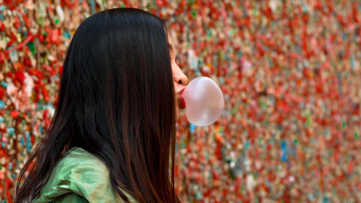 Woman blowing bubblegum bubble near a colorful gum wall, illustrating bizarre laws including a Norwegian town ban on death.