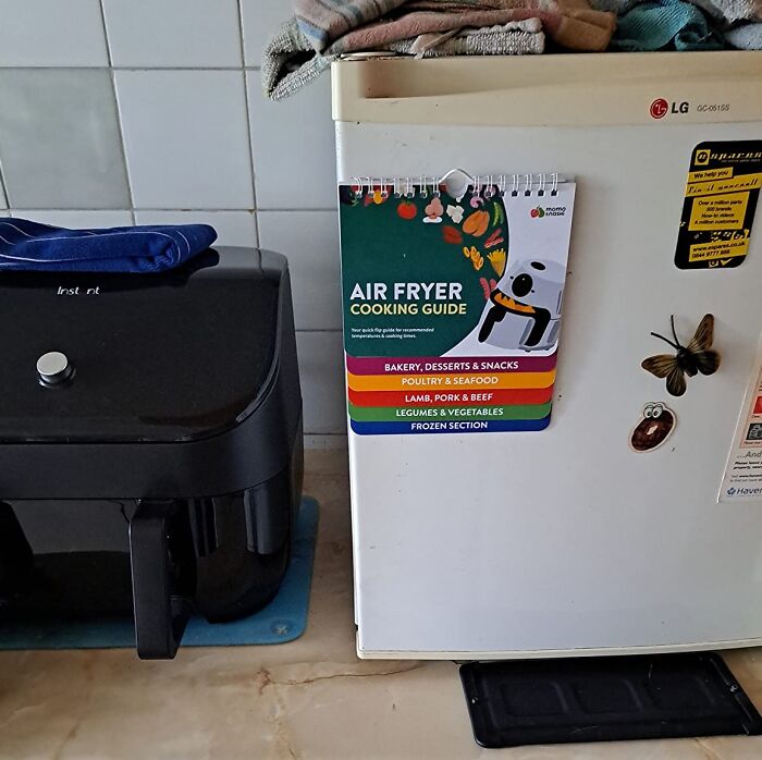 Air fryer cooking guide hanging on fridge next to black air fryer in home kitchen setting with towels on top.