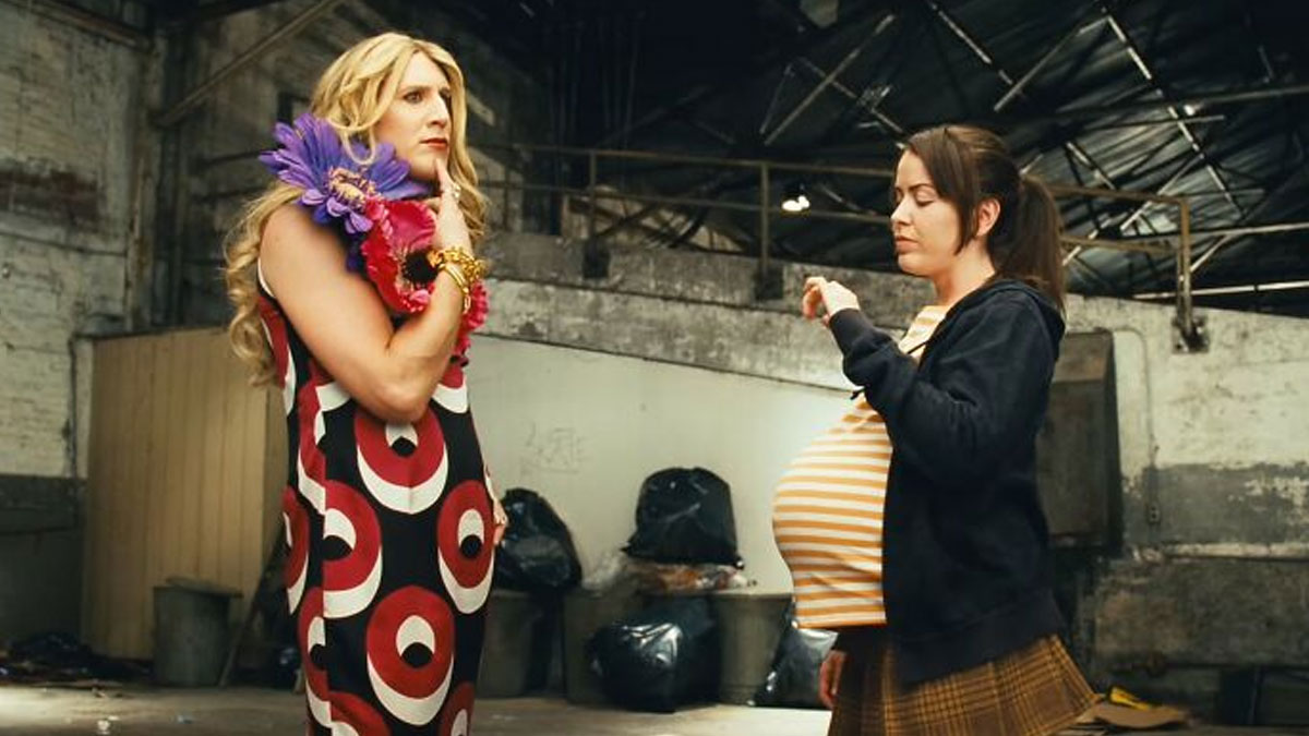 Scene from one of the worst movies featuring a man in a dress and a pregnant woman facing each other in a warehouse setting.