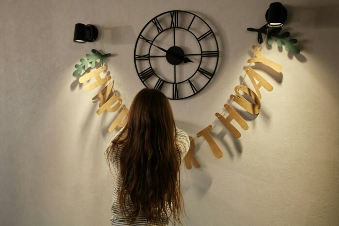 Child with long hair decorating a wall with birthday banner under a large clock, illustrating communicating calmly.
