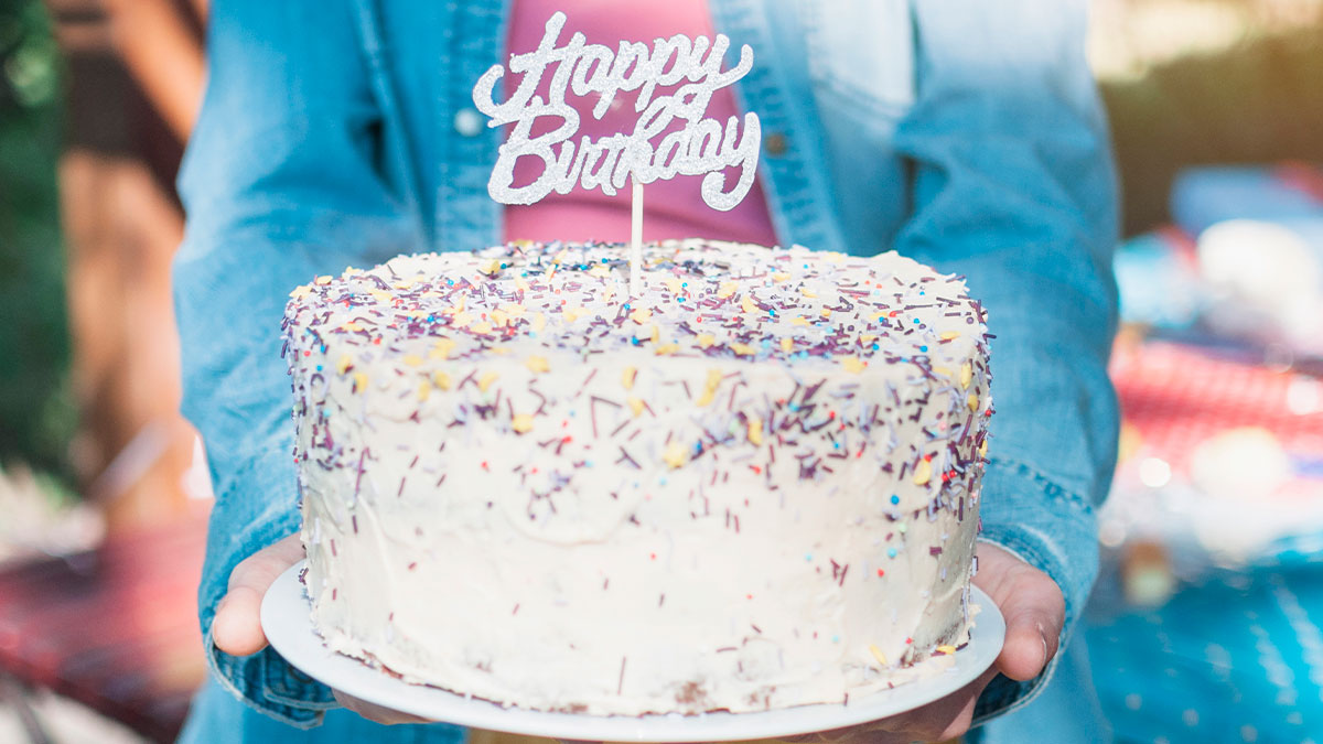 Person holding a birthday cake with white frosting and colorful sprinkles, office introvert receiving unwanted celebration.