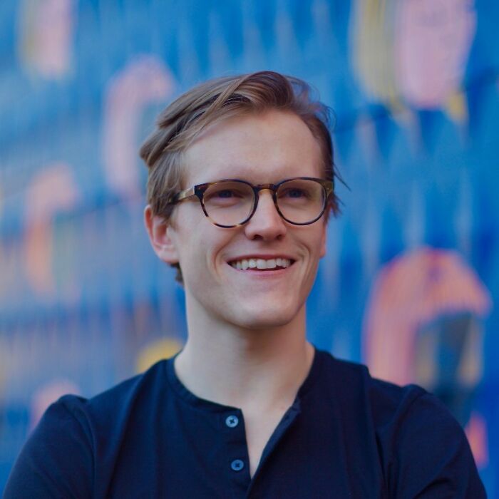Young man wearing glasses and a black shirt, smiling confidently in front of a colorful blue and orange blurred background. Young man wearing glasses and a black shirt, smiling confidently in front of a colorful blue and orange blurred background.