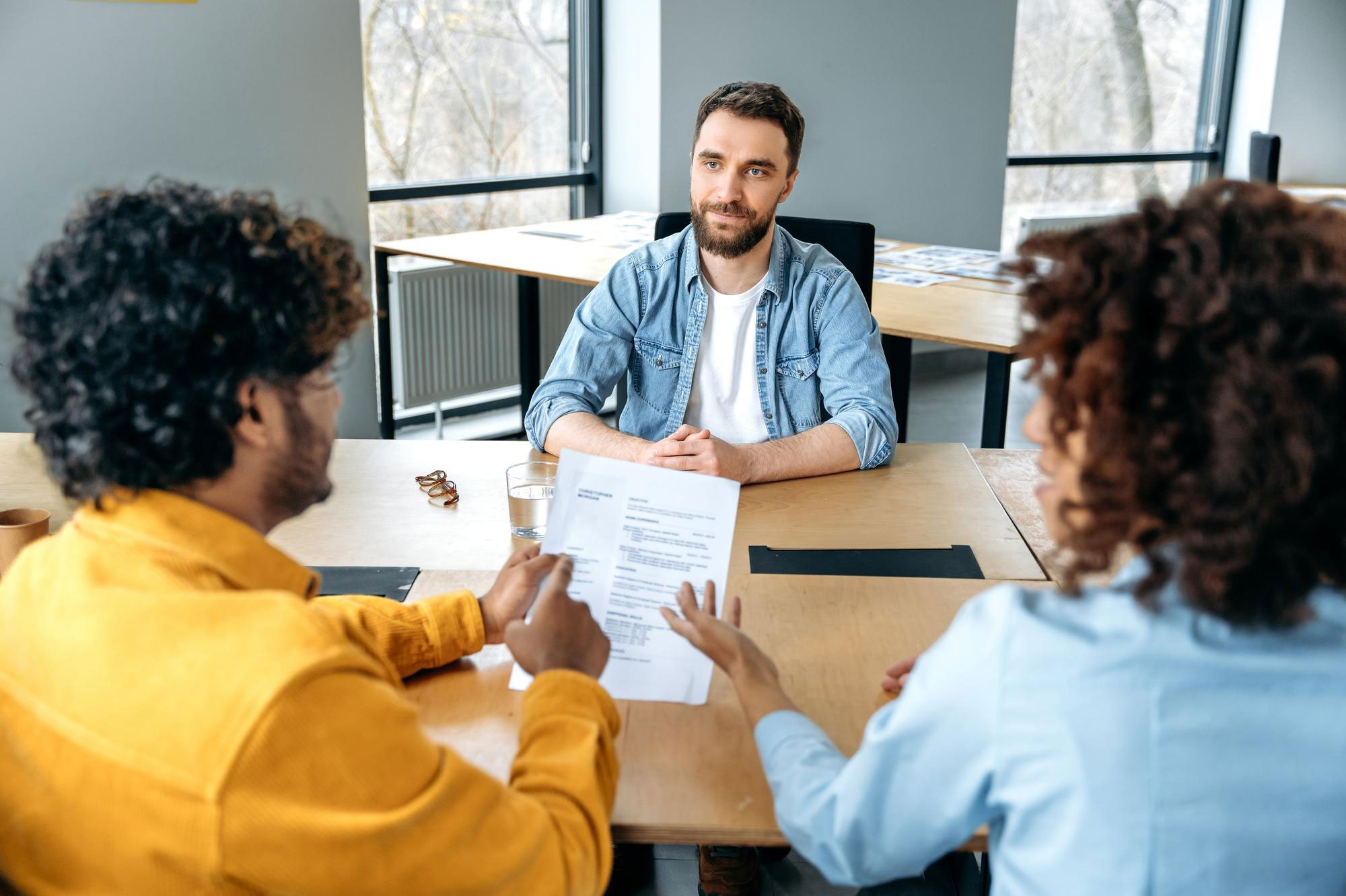 Man in denim shirt sitting in an interview setting with two people reviewing a resume discussing career and job decisions. Man in denim shirt sitting in an interview setting with two people reviewing a resume discussing career and job decisions.