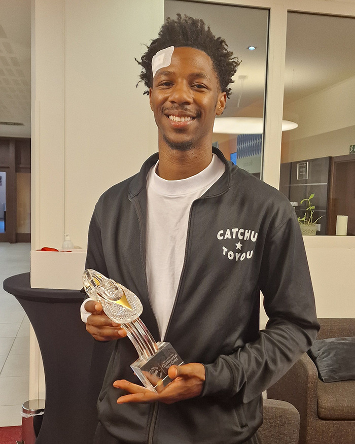 Viral sprinter smiling and holding a crystal trophy while wearing a black jacket indoors after race success. Viral sprinter smiling and holding a crystal trophy while wearing a black jacket indoors after race success.