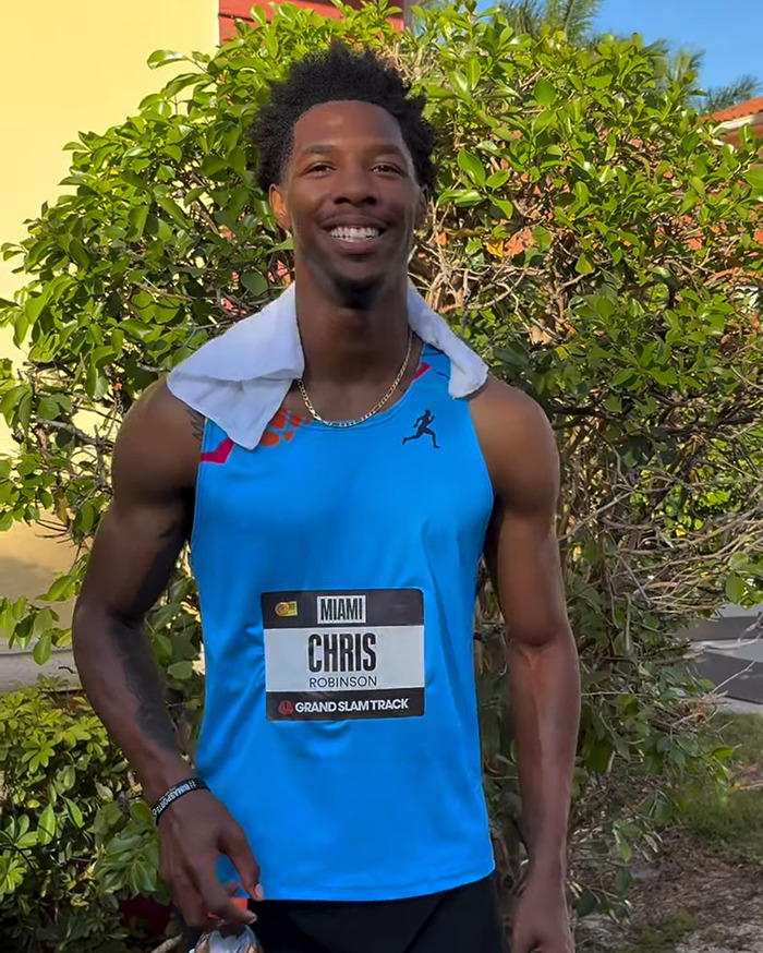 Viral sprinter Chris Robinson smiling outdoors after track event, wearing race bib and blue athletic tank top. Viral sprinter Chris Robinson smiling outdoors after track event, wearing race bib and blue athletic tank top.