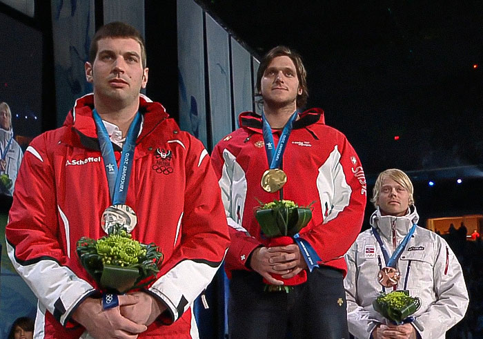 Three Olympic medalist skiers wearing medals and holding flowers during a podium ceremony at a nighttime event. Three Olympic medalist skiers wearing medals and holding flowers during a podium ceremony at a nighttime event.