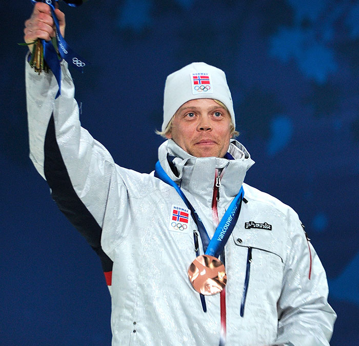 Norwegian Olympic medalist skier wearing white jacket and hat, holding up medal in a celebratory pose. Norwegian Olympic medalist skier wearing white jacket and hat, holding up medal in a celebratory pose.