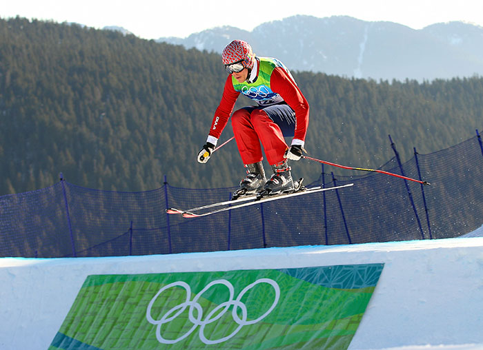 Olympic medalist skier in mid-air during a jump at a snowy mountain slope with Olympic rings banner below. Olympic medalist skier in mid-air during a jump at a snowy mountain slope with Olympic rings banner below.