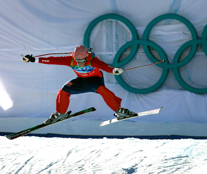 Olympic medalist skier wearing red outfit performing a jump with Olympic rings in background on a snowy slope. Olympic medalist skier wearing red outfit performing a jump with Olympic rings in background on a snowy slope.
