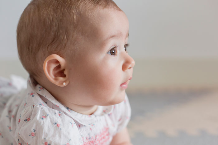 Baby with gold stud earrings after unlicensed ear piercing during a family visit, mom visibly upset in a home setting. Baby with gold stud earrings after unlicensed ear piercing during a family visit, mom visibly upset in a home setting.