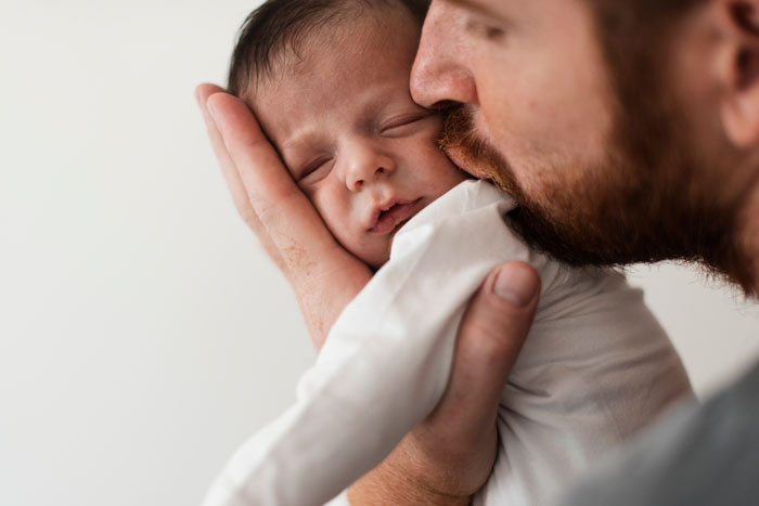 Father carrying infant by the arms while gently wiping the baby's face with a dishrag, unaware of wife's concern. Father carrying infant by the arms while gently wiping the baby's face with a dishrag, unaware of wife's concern.