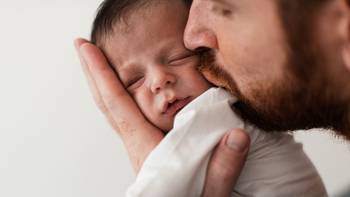 Father tenderly holding and kissing a sleeping infant close to his face, showing gentle infant care and bonding.