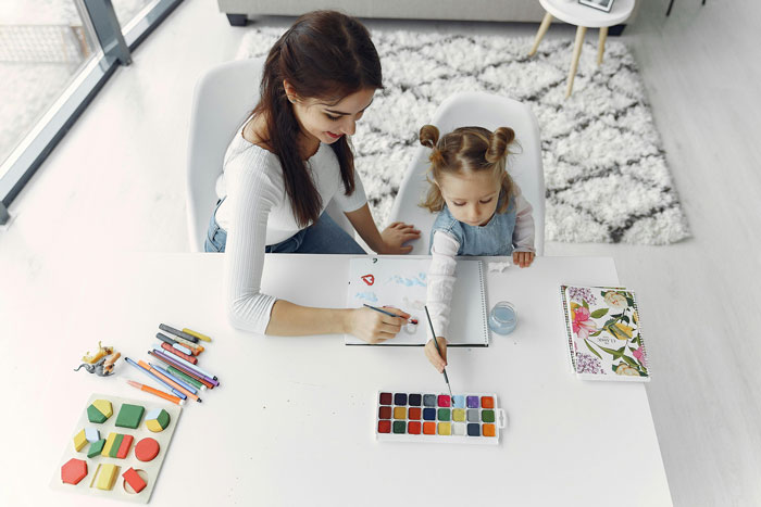 Woman babysitting young girl with paints and crayons at white table in bright living room Woman babysitting young girl with paints and crayons at white table in bright living room