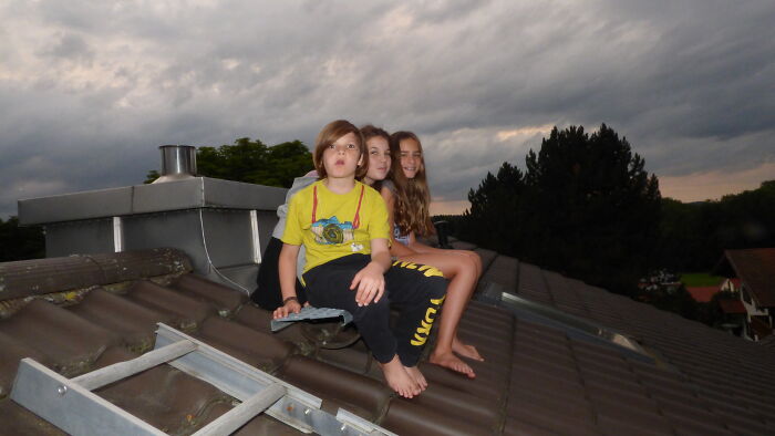 Three children sitting barefoot on a rooftop at dusk, evoking a creepy babysitter story atmosphere under darkening skies.