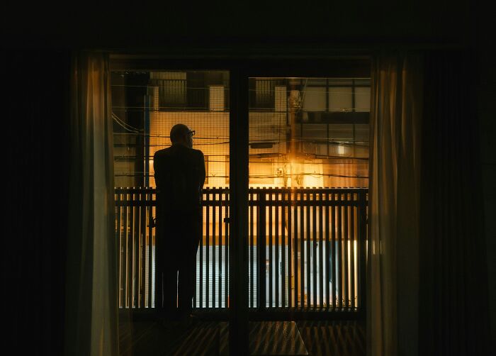 Silhouette of a person standing inside a dimly lit room looking out a window, evoking creepy babysitter story vibes.