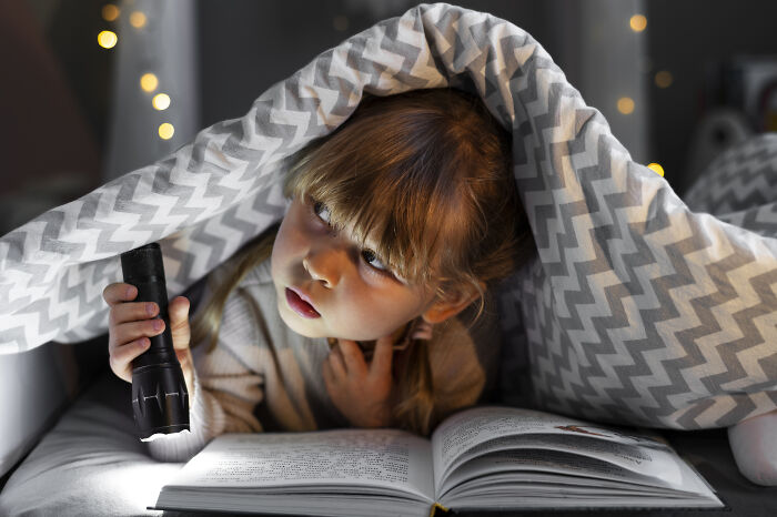 Child babysitter reading a book under a blanket with a flashlight, creating a creepy babysitter story atmosphere at night.