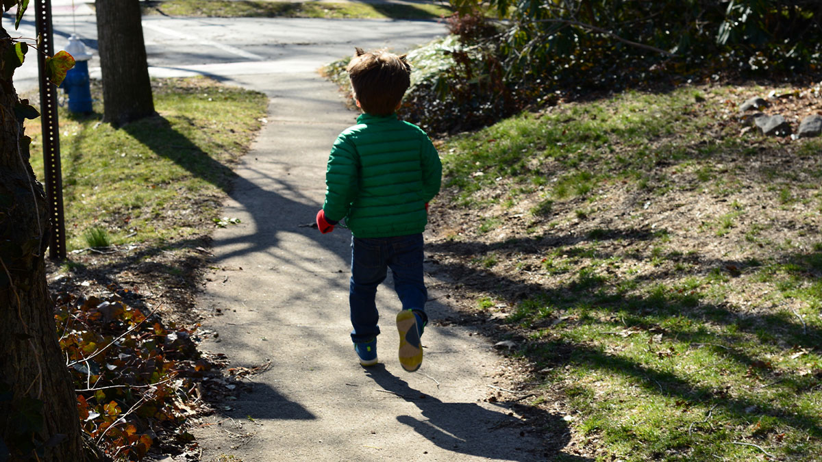 Young child in green jacket running alone on a sunny sidewalk, evoking creepy babysitter stories atmosphere