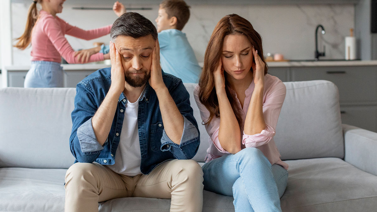Young adults stressed on couch while noisy children play in background, depicting challenges of babysitting multiple siblings.