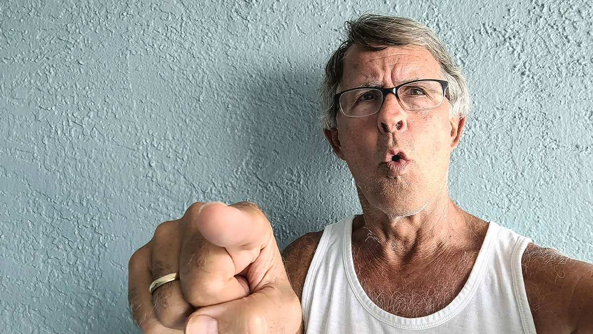 Angry older man wearing glasses and white tank top pointing aggressively at the camera inside a barbershop setting.
