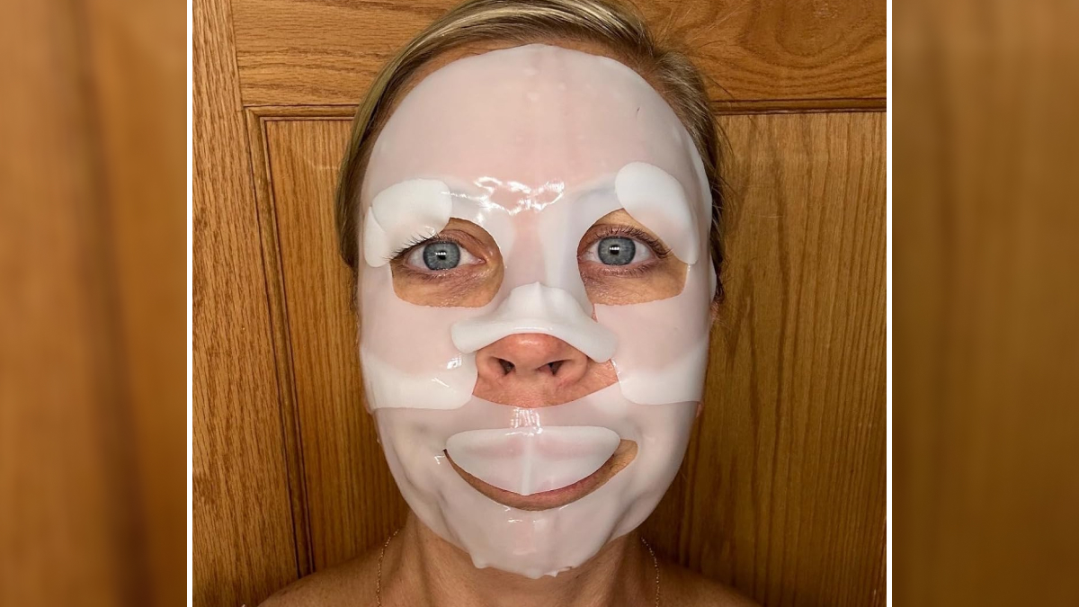 Woman wearing a white facial sheet mask standing in front of a wooden door during skincare routine for Prime Day deals.