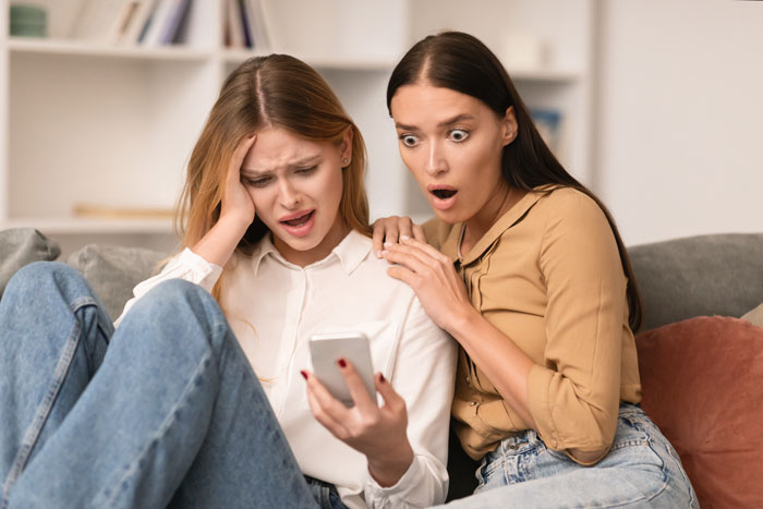 Two young women, one upset and the other shocked, looking at a smartphone, reflecting influencer drama and studio party conflict. Two young women, one upset and the other shocked, looking at a smartphone, reflecting influencer drama and studio party conflict.
